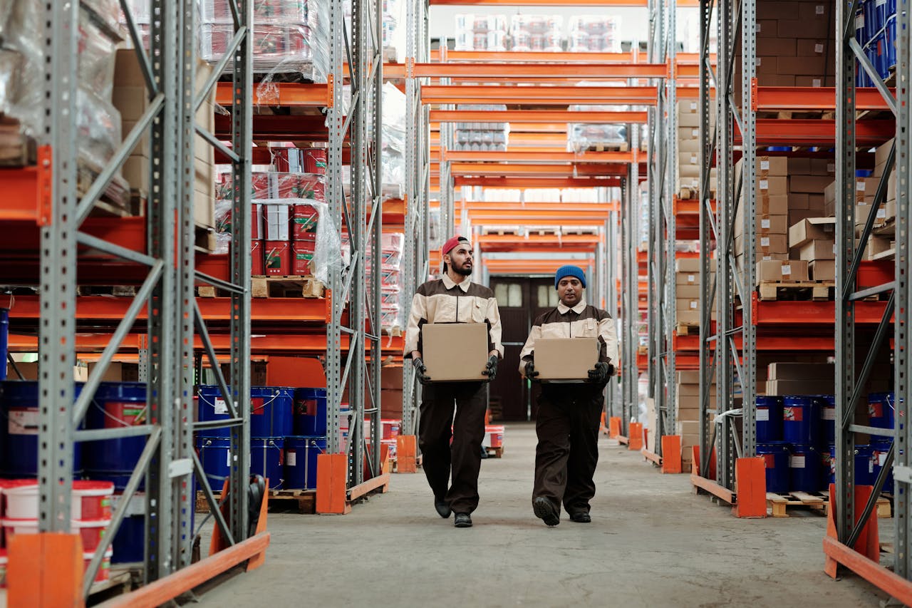 Two workers carrying boxes walk between metal shelves in a spacious warehouse.